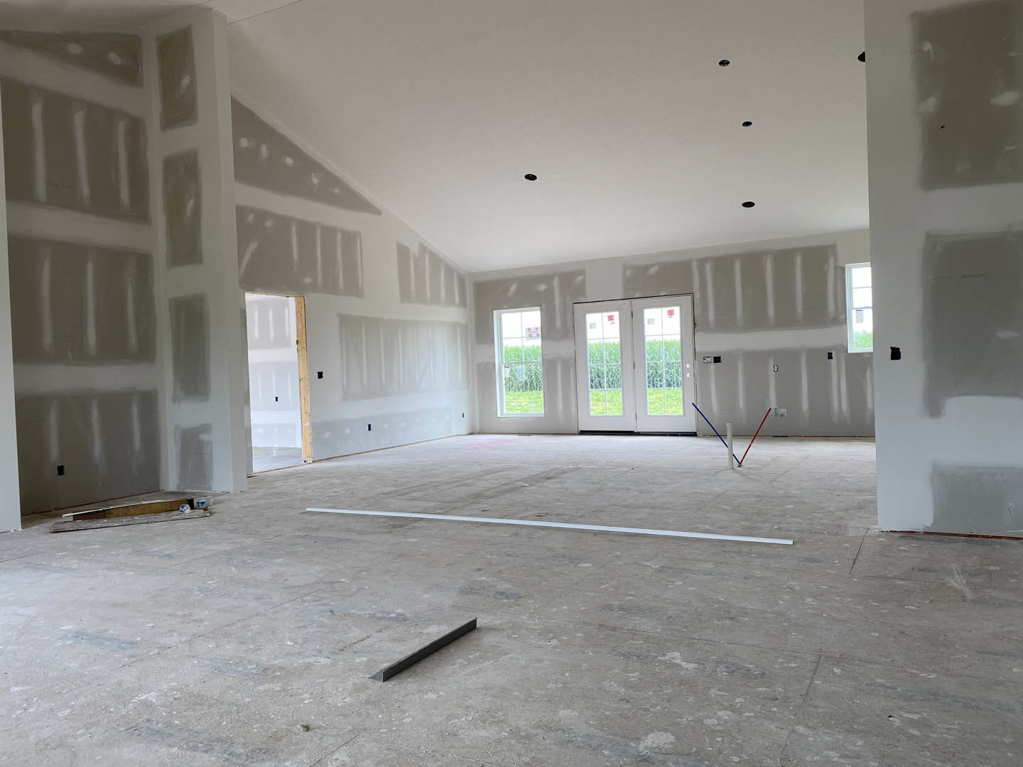 White-walled room with white ceiling, concrete floor featuring a metal strip, double glass-paneled doors, two windows—one with a green plant visible outside, the other displaying a