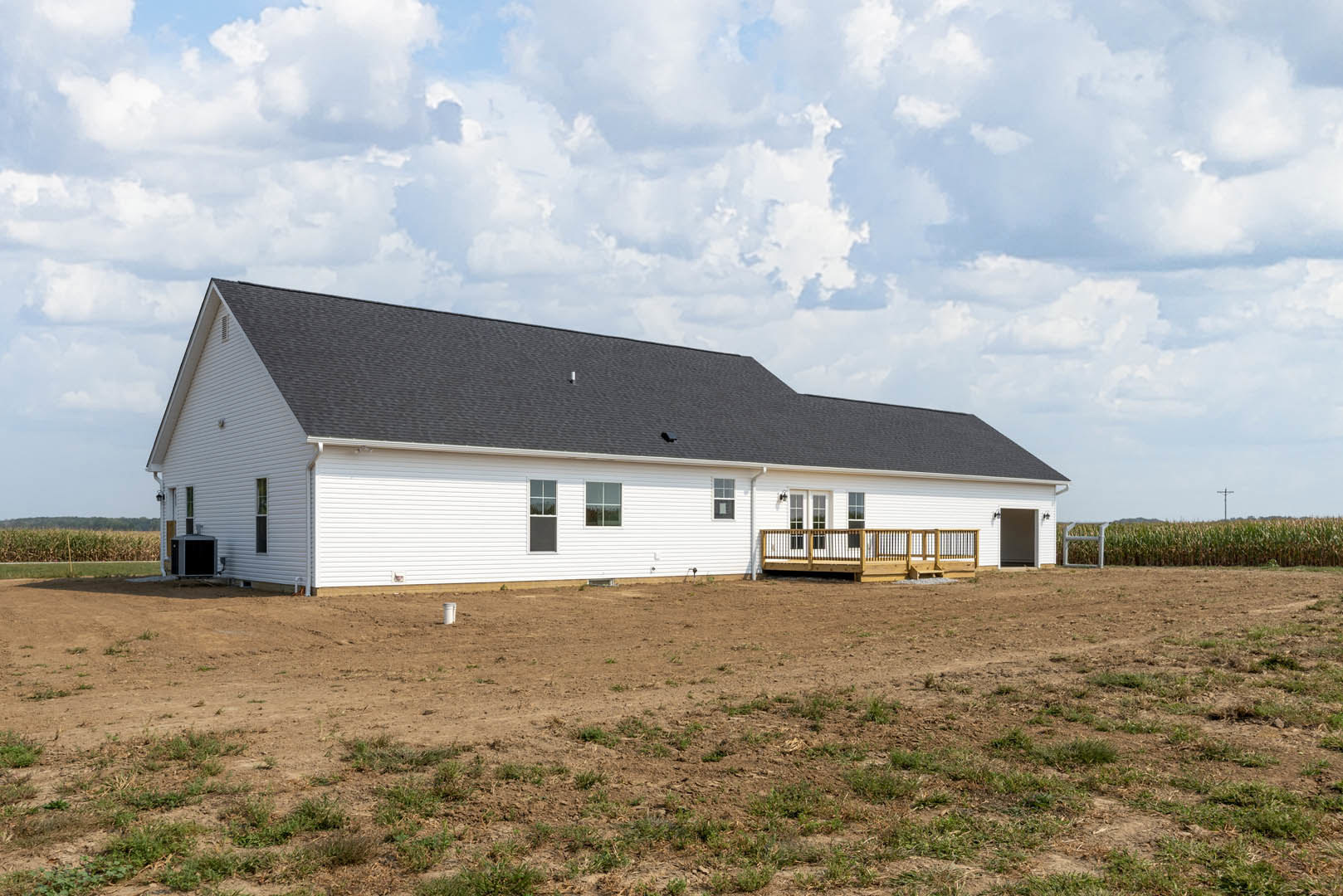 White house with black roof, wooden deck and railing, surrounded by large dirt field with patches of grass and a fence under cloudy sky
