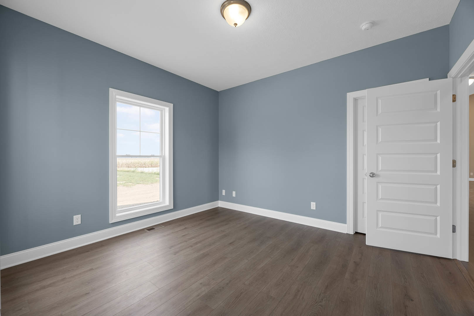 Wood flooring with white baseboards, white door with silver handle, ceiling light fixture, window overlooking a field and dirt road