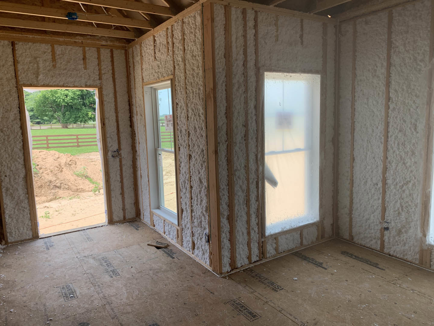 Unfinished room with exposed wood framing, insulated wall, white curtained window showing outdoor fence and trees, door to the right, construction materials visible