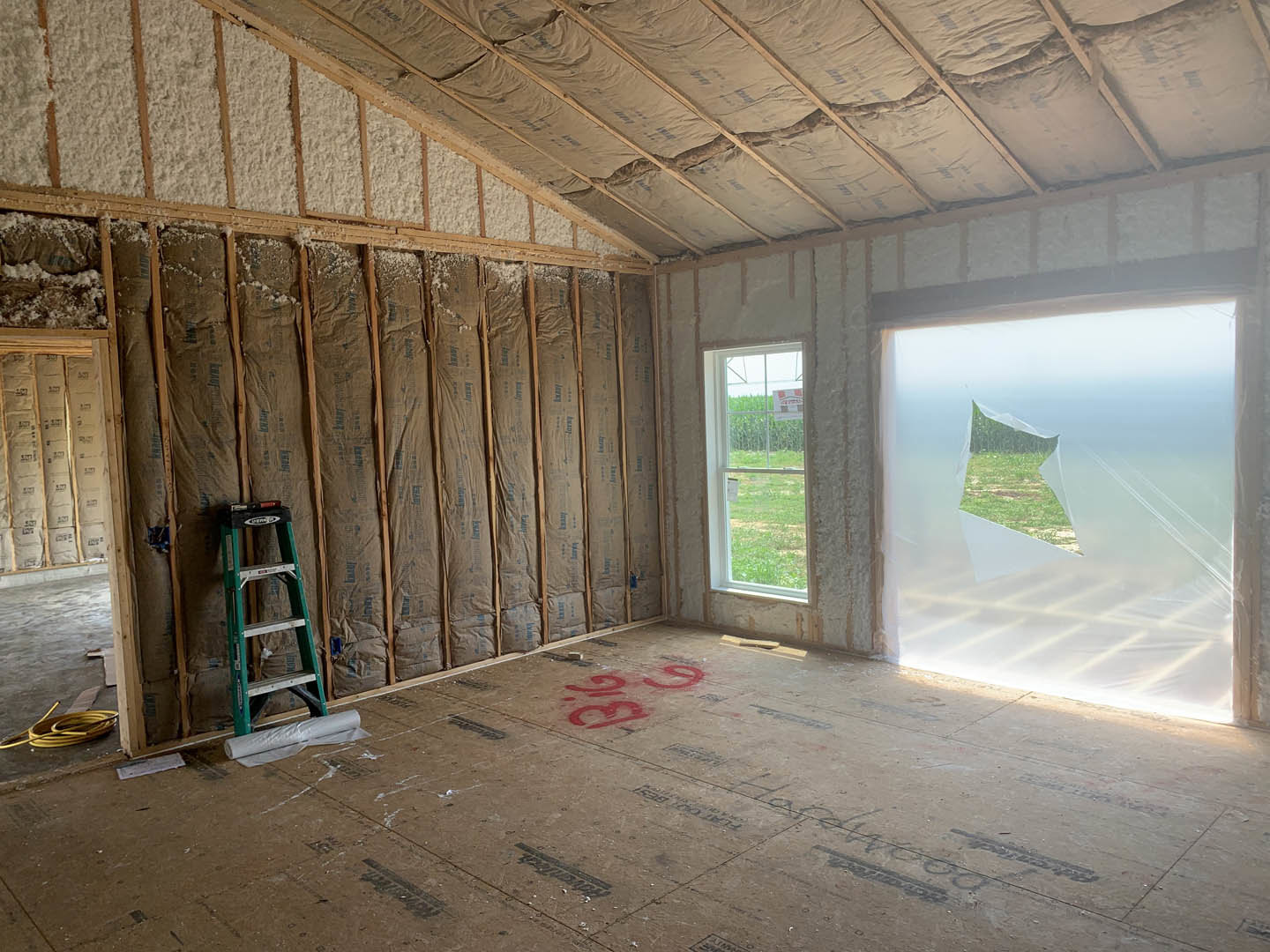 Unfinished room with exposed ceiling insulation, concrete floor marked with red numbers, wooden beams, ladder leaning against wall, window letting in natural light, and blocks of