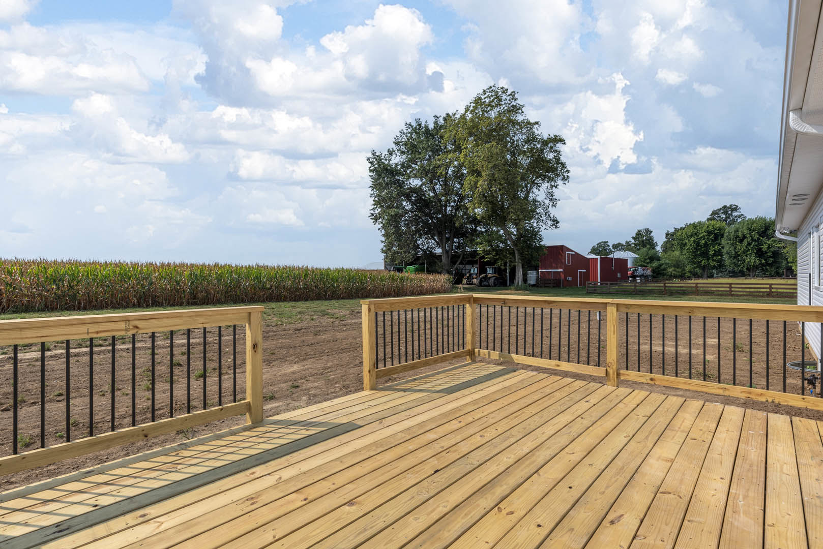 Wooden deck with black metal railings, fenced perimeter, overlooking a cornfield and surrounded by trees under a cloudy sky
