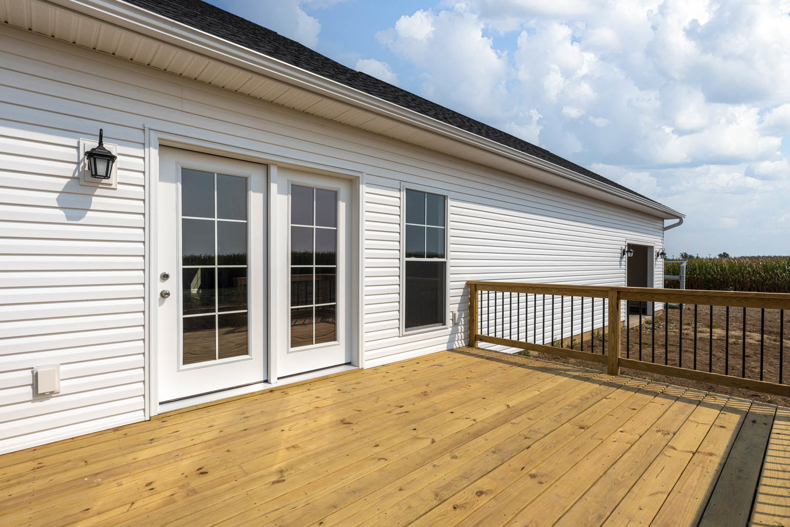 Wooden deck with black metal railing, white siding exterior, white-framed window, white door, and black wall-mounted light fixture