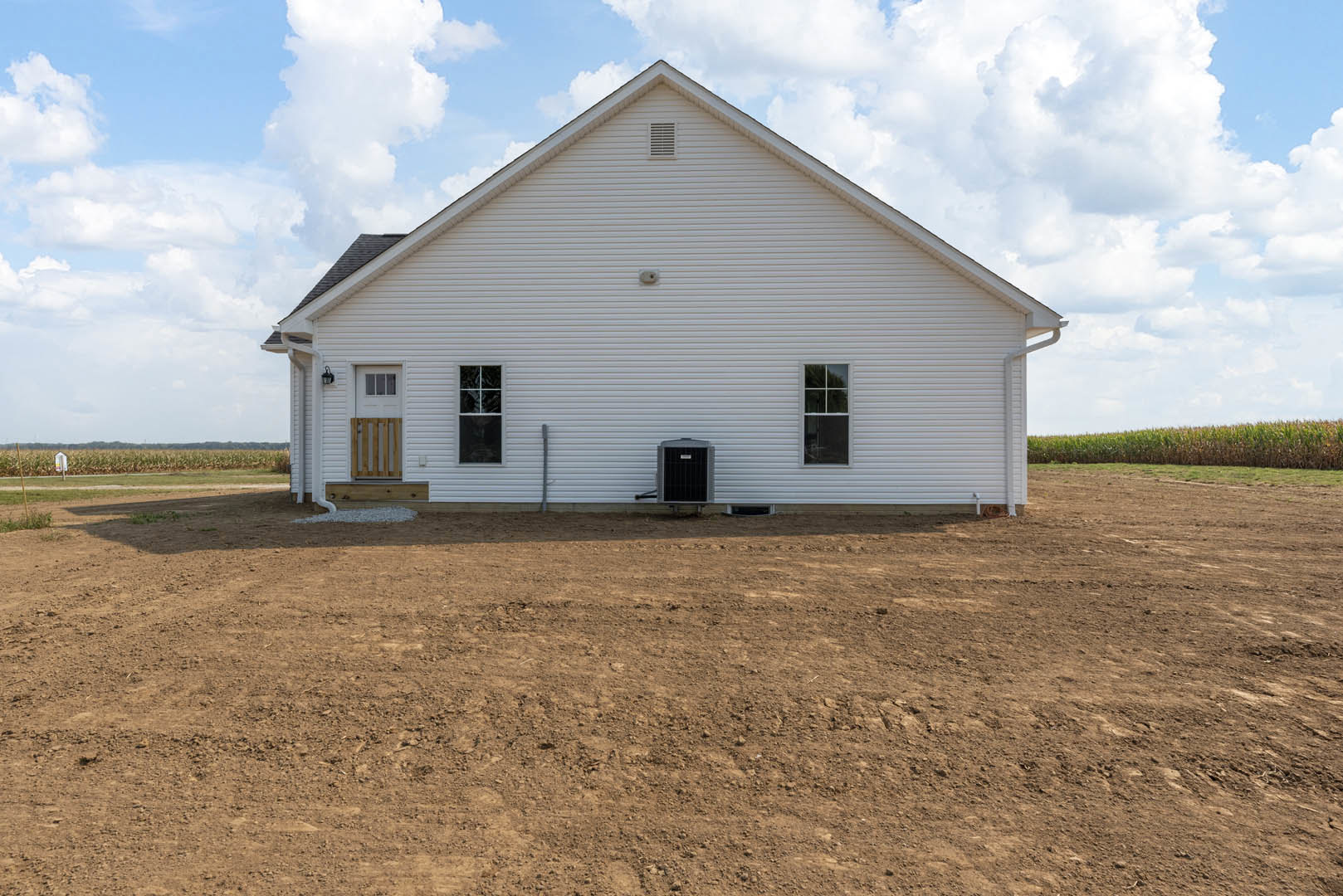 Single-story house with white-framed window, black metal door, and adjacent dirt field under cloudy sky