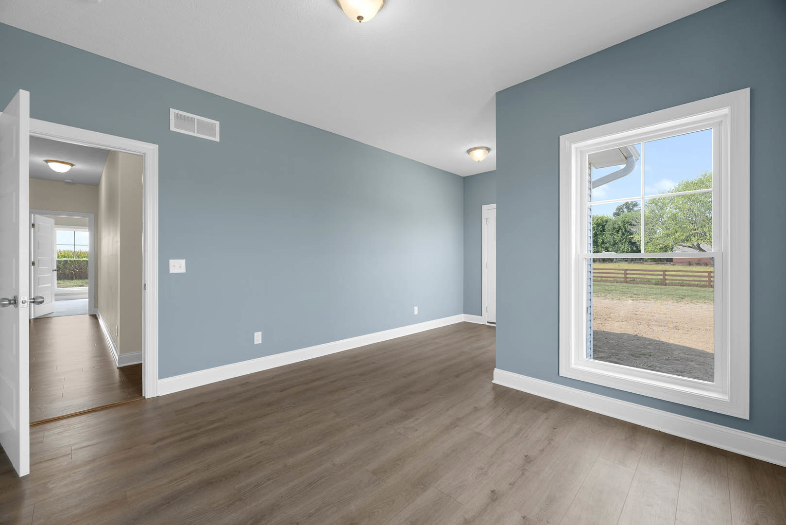 Sunlit room with hardwood flooring, large window overlooking fenced yard and trees, white plaster walls, ceiling light fixture, wall vent, and decorative molding.