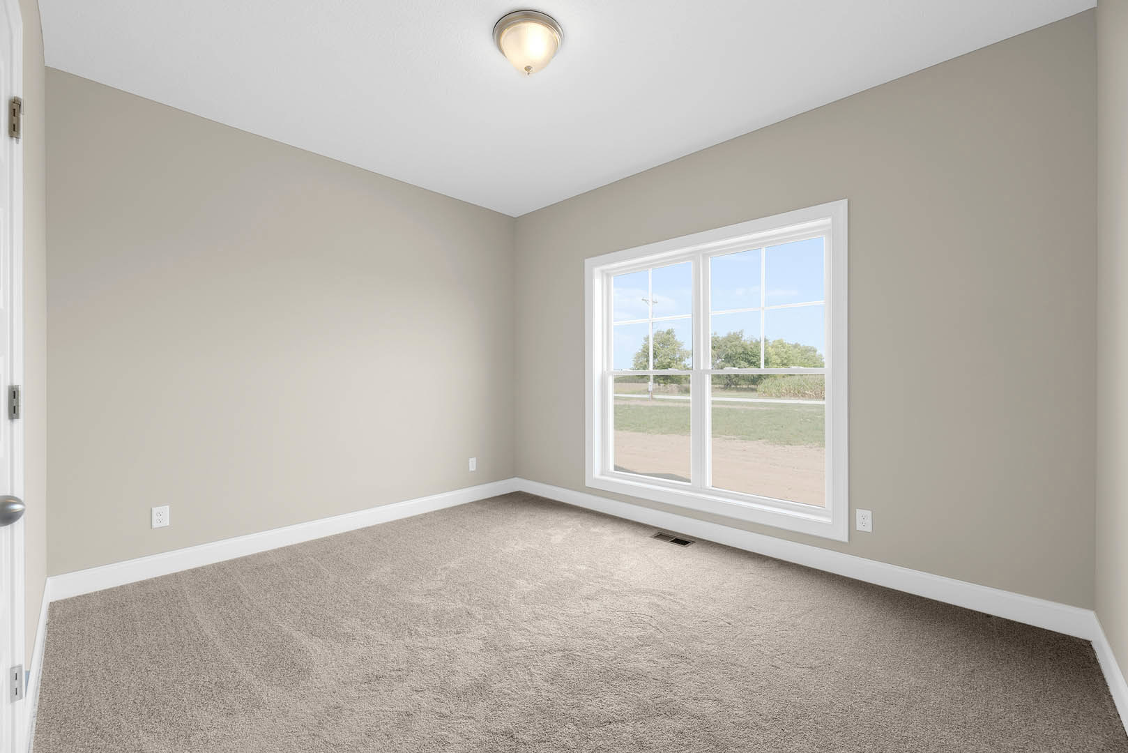 Neutral-toned carpeted room with white walls, large window overlooking field and trees, ceiling-mounted light fixture, and close-up of brushed metal door handle