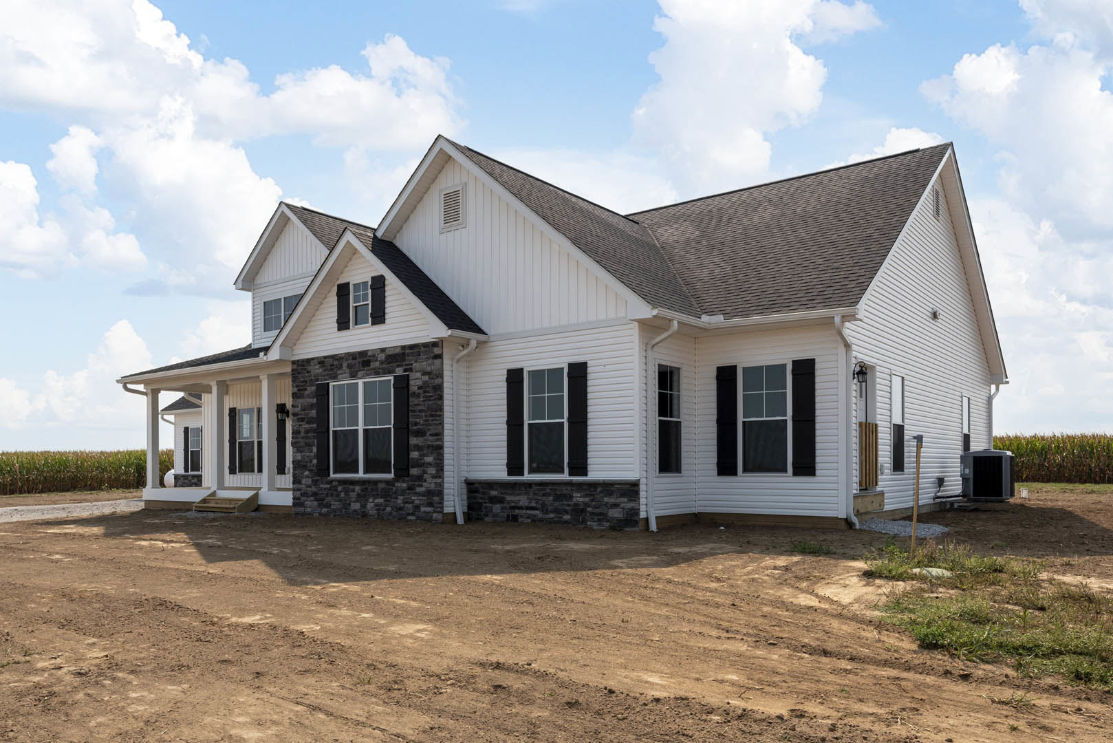Partially built house with white framed windows, black shutters, exposed dirt yard, and large black utility box under clear blue sky