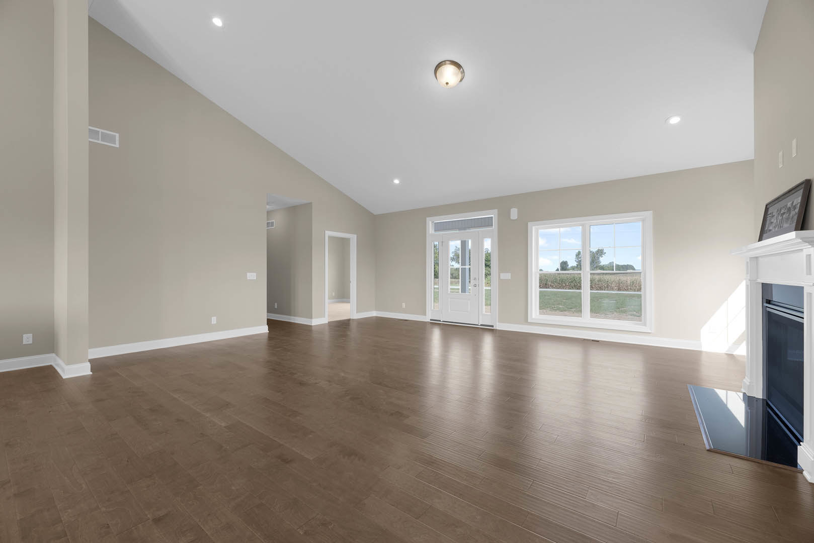 Spacious room with hardwood flooring, white glass-paneled door, ceiling light fixture, large window overlooking a grassy field, and framed sign on wall