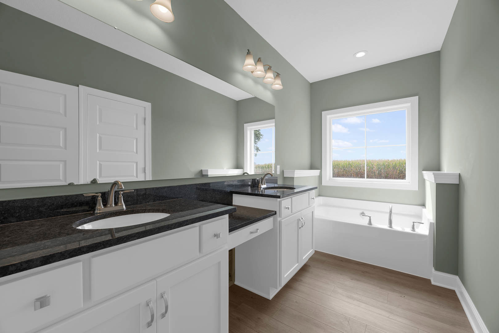Bathroom featuring white cabinetry, black countertops, chrome faucet, green accent wall, white door, window overlooking grassy field, and multi-bulb light fixture.