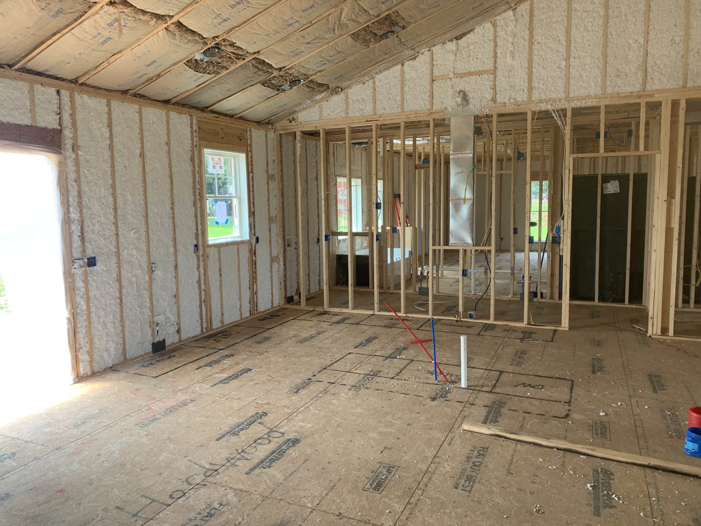 Spacious room with white plaster walls, exposed ceiling beams, large window letting in natural light, and unfinished floor featuring blue and red poles.