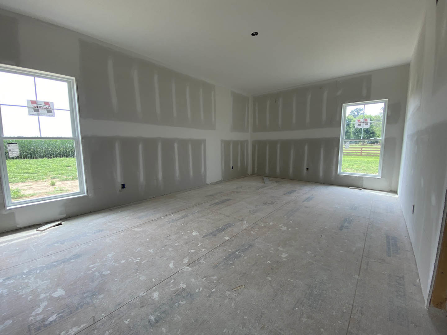 Bright room with white plaster walls, large windows, and unfinished flooring; window features a white sign.