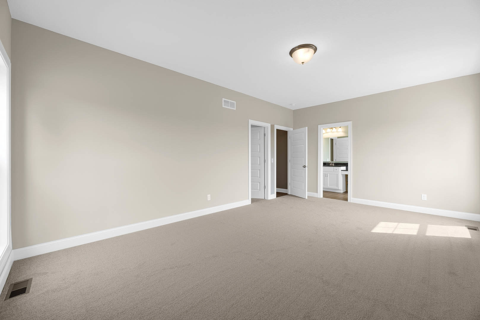 Beige carpeted room with white walls, open white door with silver handle, white door frame, ceiling light fixture, partial view into bathroom with mirror