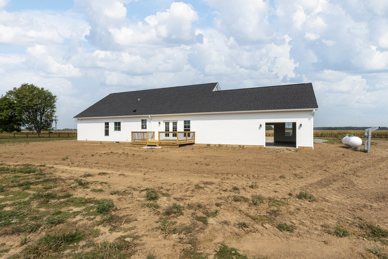 White siding house with expansive wooden porch, black metal railing, grassy yard, leafy tree, and gabled roof under partly cloudy sky