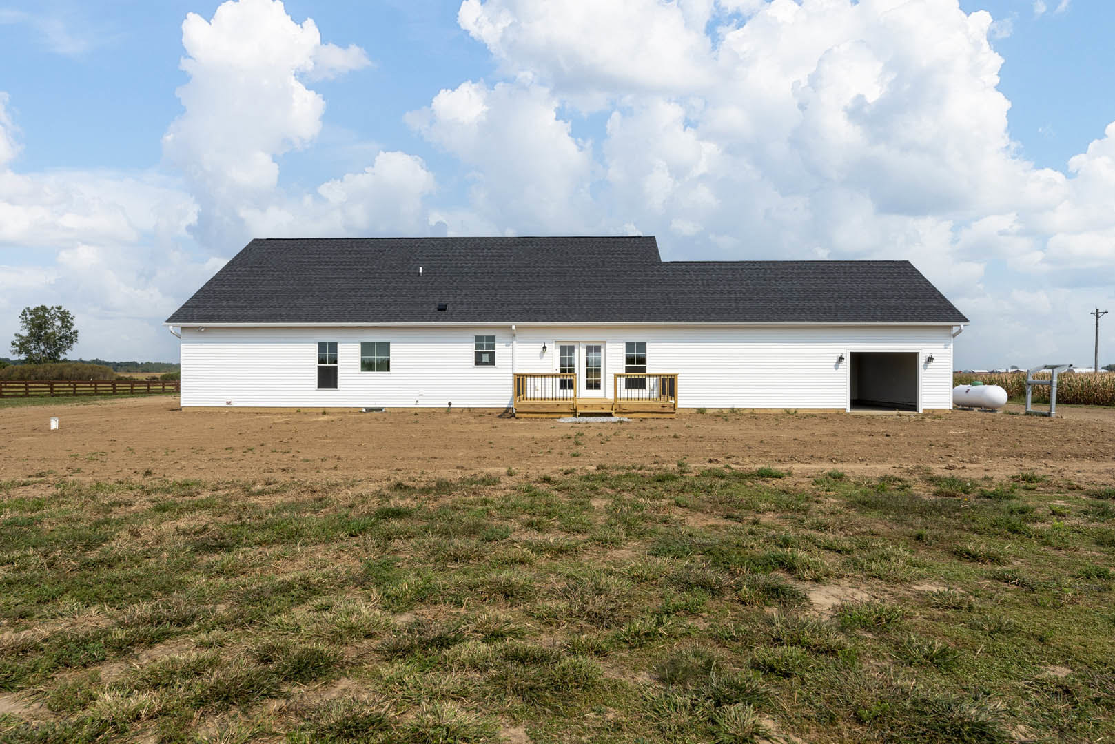 White house with black roof, wooden porch and railings, surrounded by large grassy field, tree in foreground, clouds in blue sky