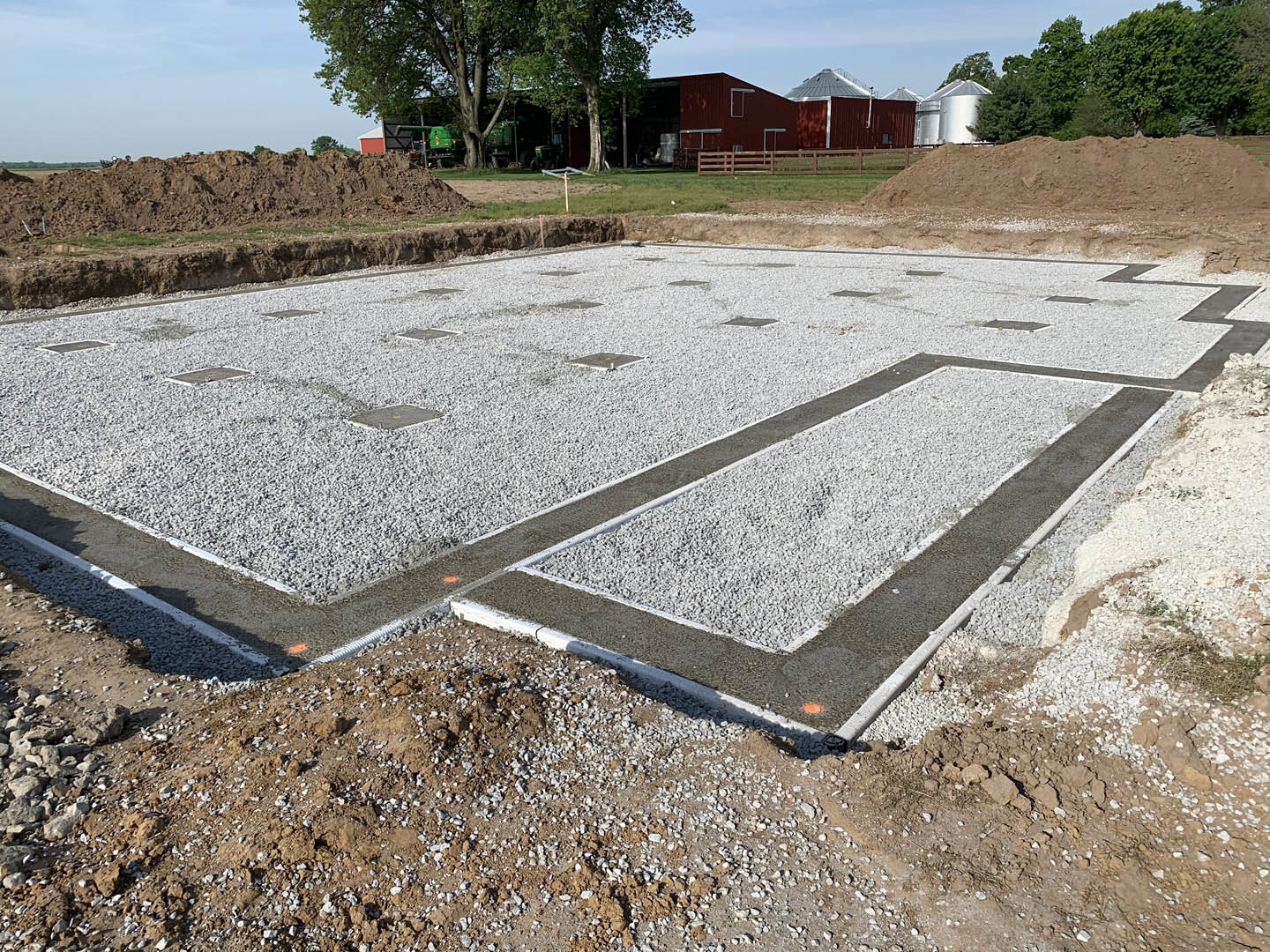Concrete foundation under construction with gravel fill, surrounded by grass, large tree, tractors, silver silo, and utility pole in the background.