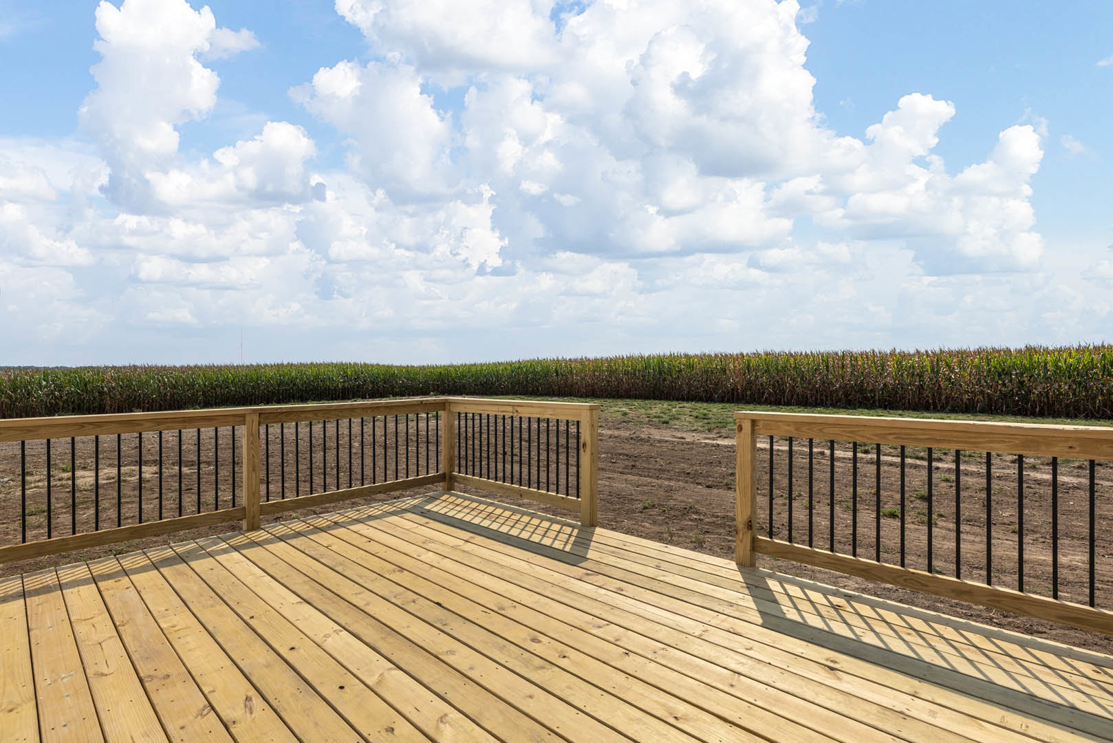 Wooden deck with black metal railing, fenced perimeter, overlooking a cornfield under a partly cloudy blue sky