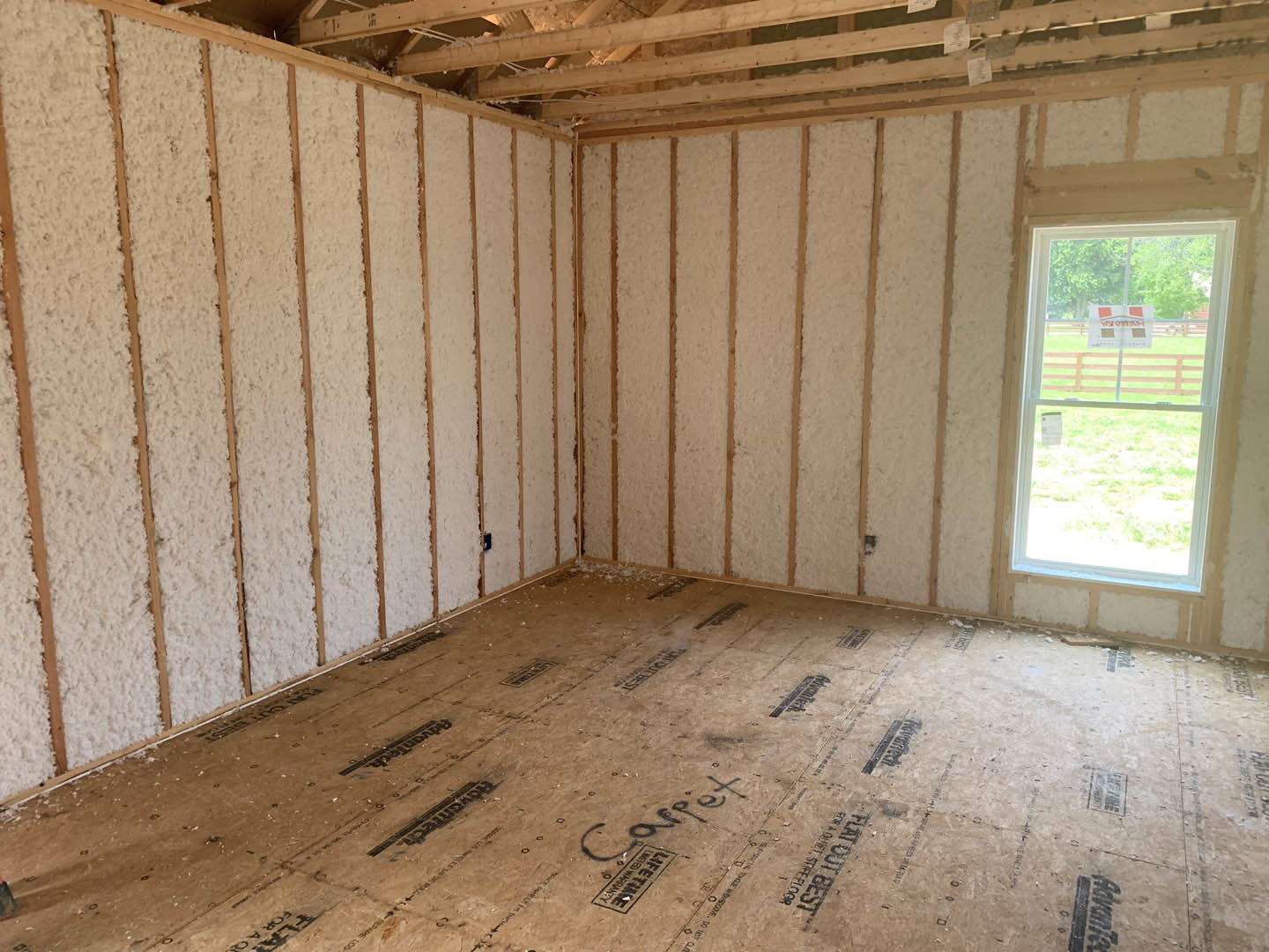 Room with unfinished wood plank walls, exposed ceiling beams, foam insulation covering the subfloor, and a window displaying a sign.