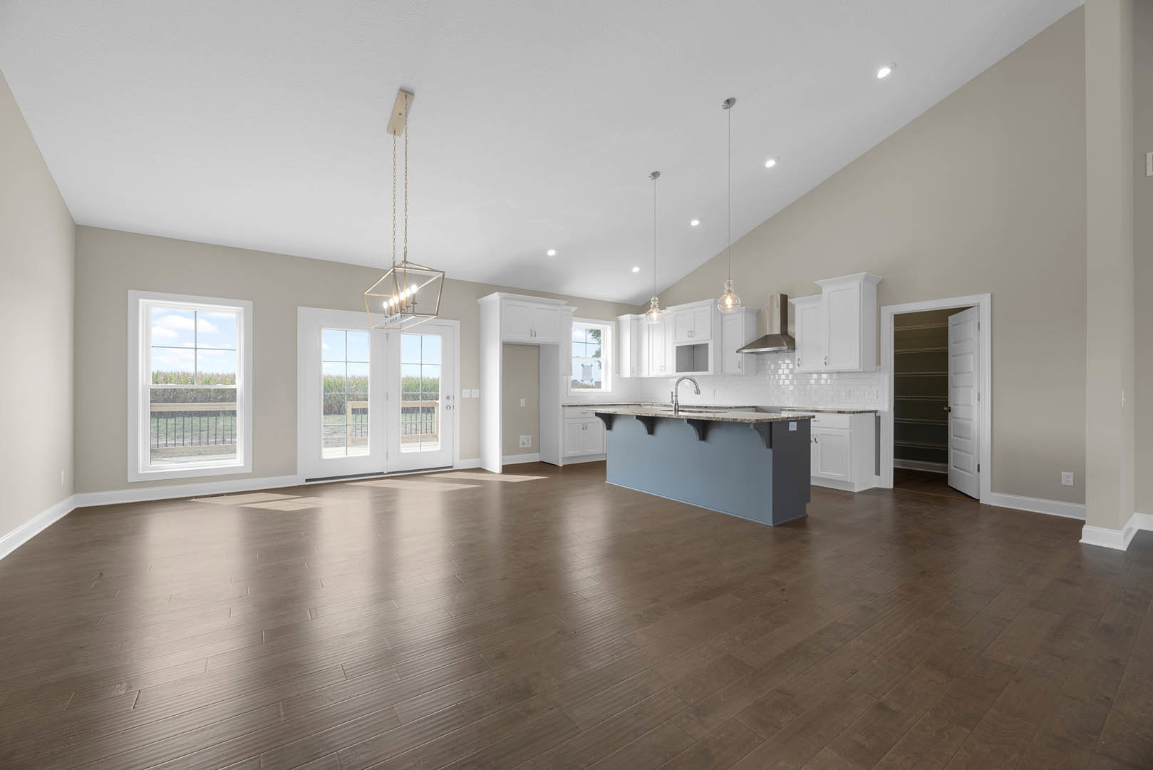 Spacious open-concept kitchen and living area featuring a central island with white countertops, hardwood floors, large windows overlooking a field, and neutral walls with white