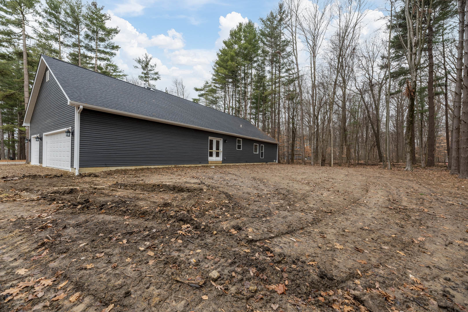 Black siding house with white glass-paneled door, dirt field covered in leaves, group of trees, blue sky with clouds in the background