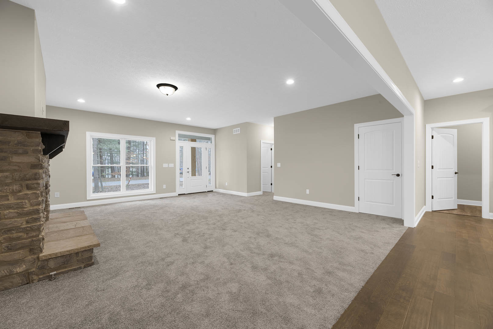 Wood staircase with natural finish, white door featuring black handle, carpeted floor, white walls, window overlooking trees, exposed brick wall with opening, white door with glass