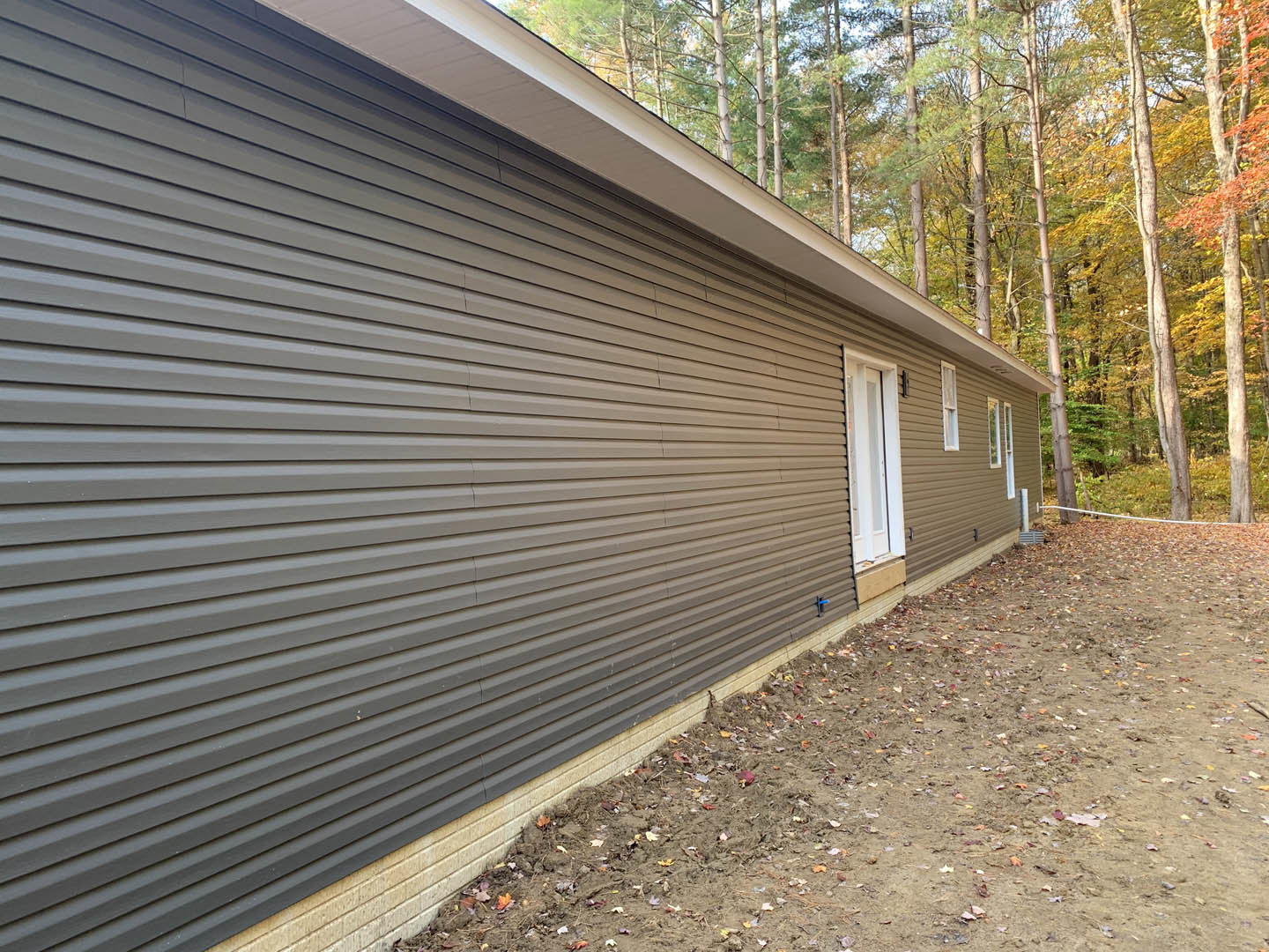 Modern house with light siding and large windows, surrounded by autumn trees, dirt path running alongside the exterior wall