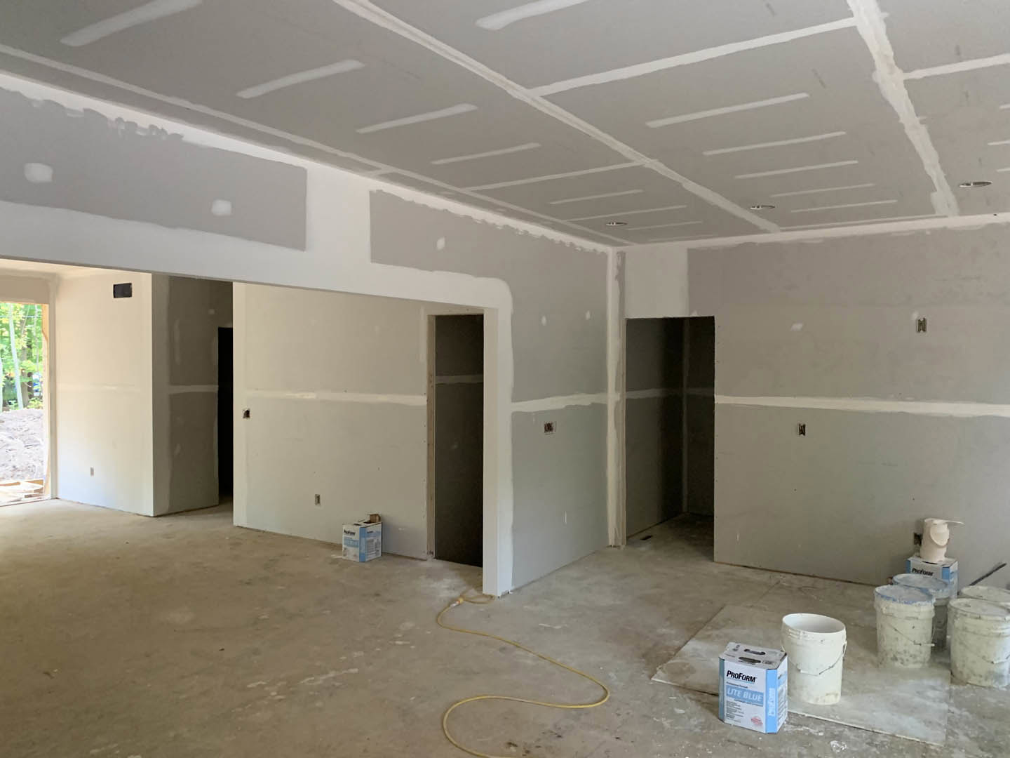 Unfurnished room with freshly painted white walls, paint buckets on unfinished concrete floor, white ceiling, close-up of door, and roll of toilet paper near wall