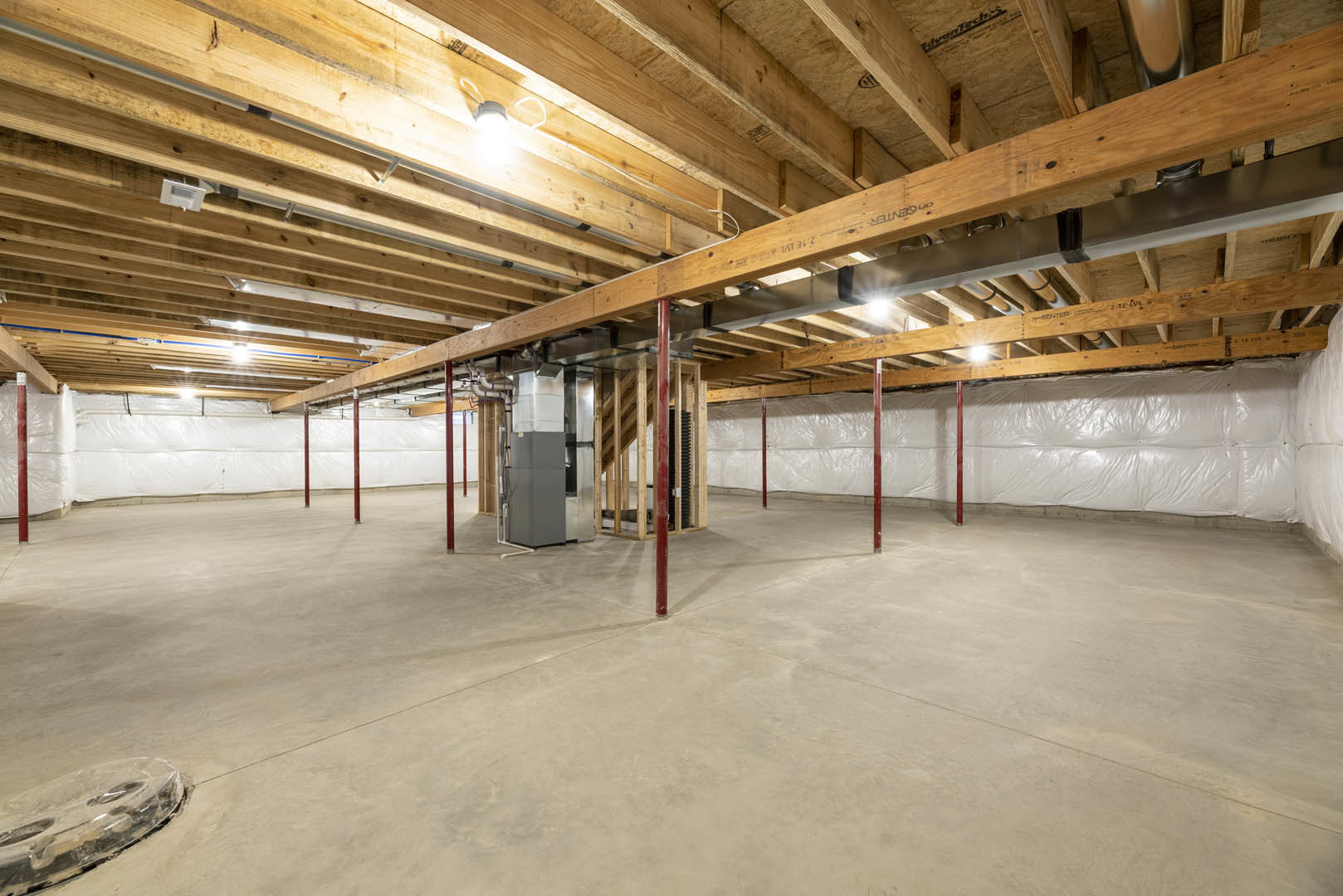 Basement room with exposed wood ceiling beams, metal pipes, concrete floor, red support pole, white rectangular window unit, and building insulation along walls