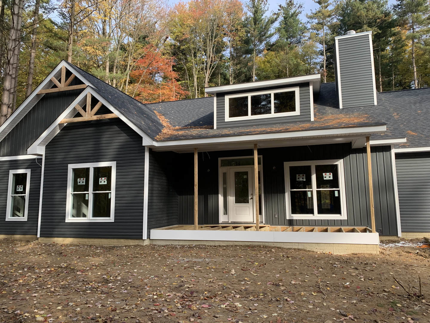 House under construction with white door featuring glass panel, window displaying stickers, dirt ground in foreground, surrounded by dense trees in background