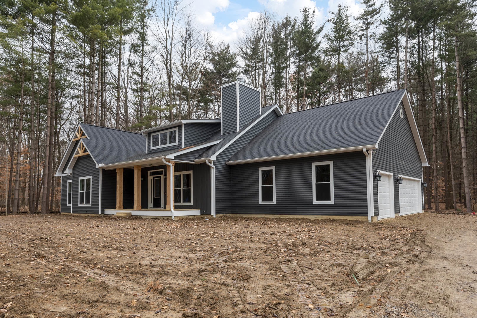 Partially built house with exposed framing, white window trim, brick chimney, dirt yard, and mature trees in the background