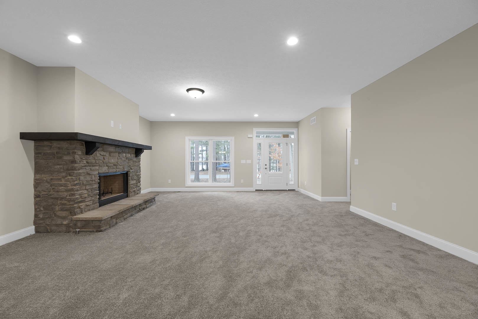 Living room with gray carpet, stone fireplace, white door with glass panes, large window showing trees outside, plaster ceiling and walls