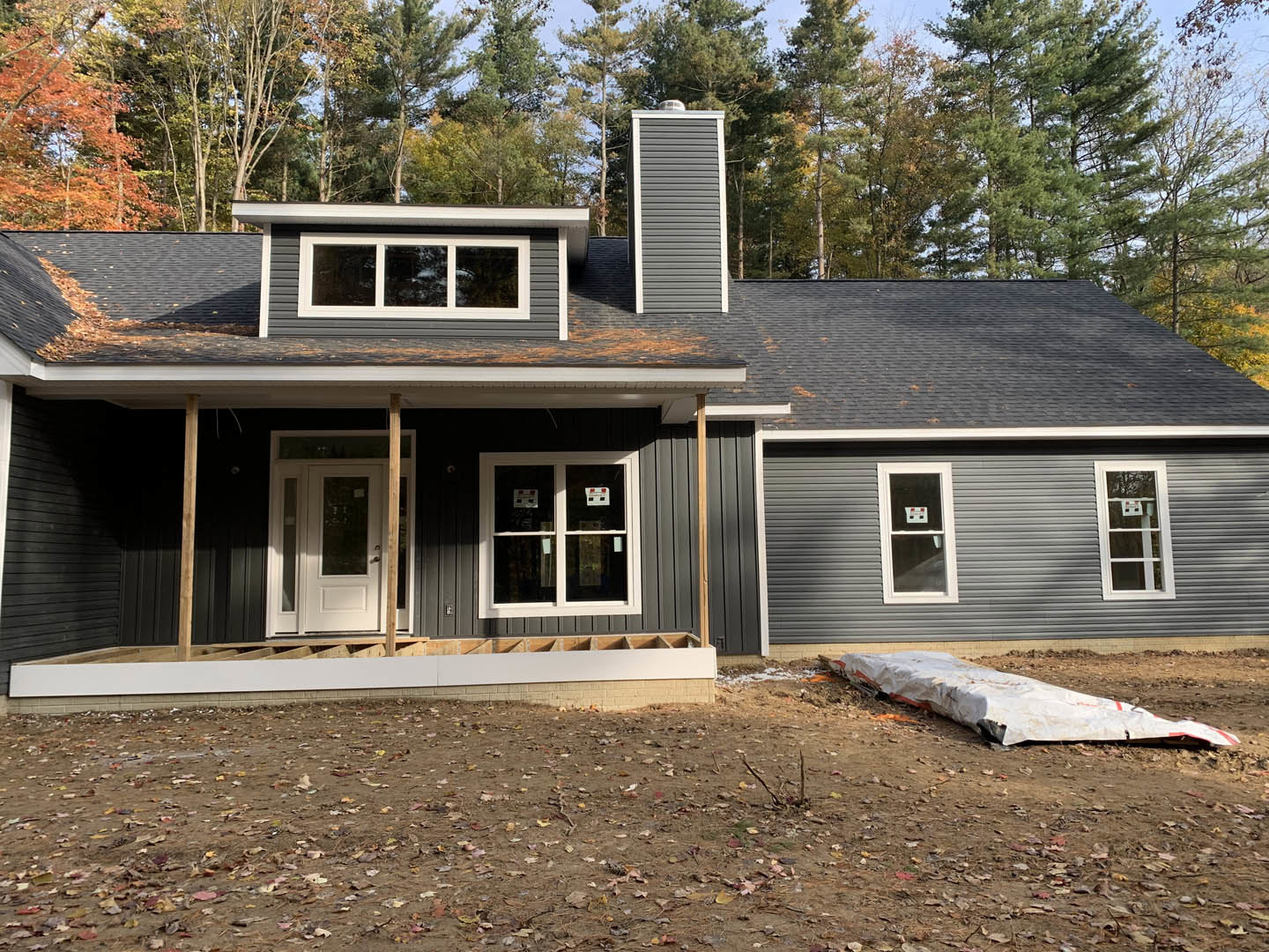 Partially built house with exposed framing, white-framed windows, white tarp spread on dirt ground, scattered leaves, and mature trees in the background