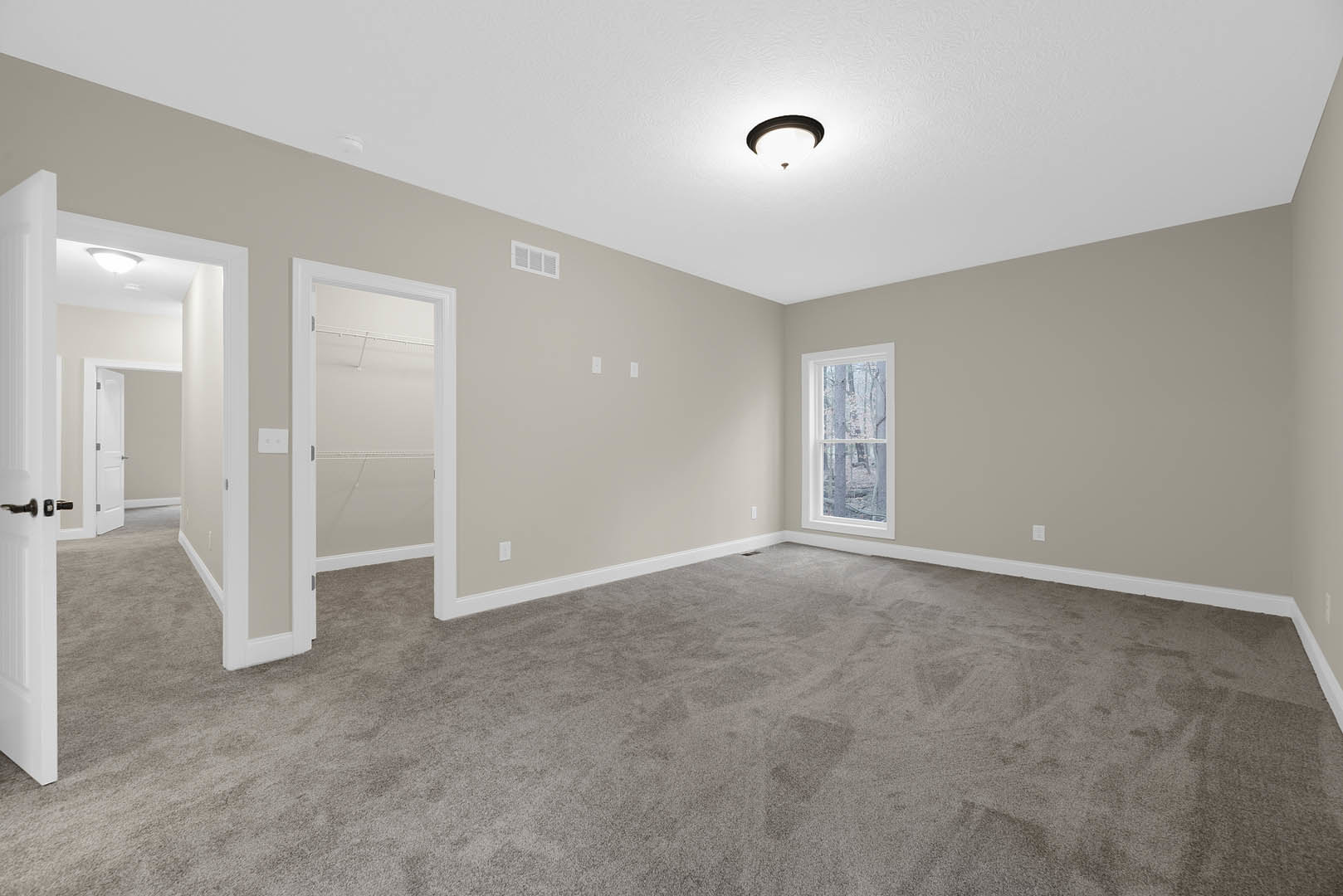 Beige-walled carpeted room with white door, window overlooking trees, and ceiling light fixture with black ring