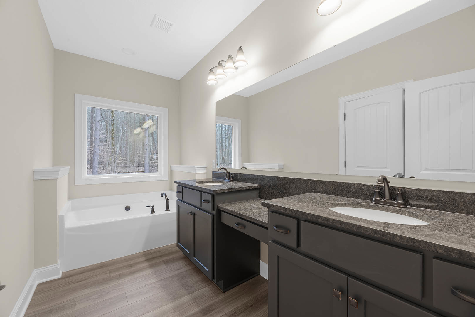 Bathroom with freestanding bathtub, white sink on stone countertop, four-bulb light fixture, large window overlooking trees, neutral tile walls, wood cabinetry, chrome faucet