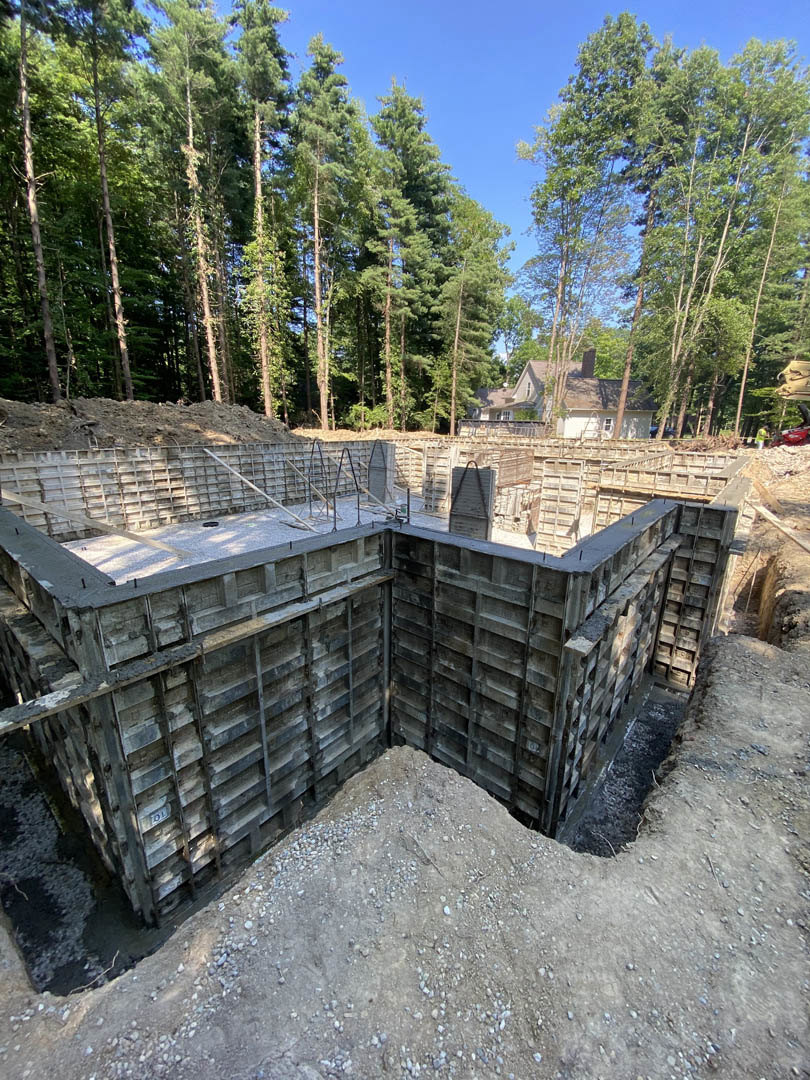 Concrete foundation and metal framing of a custom home under construction, surrounded by mature trees, with stone and wood materials visible on site.