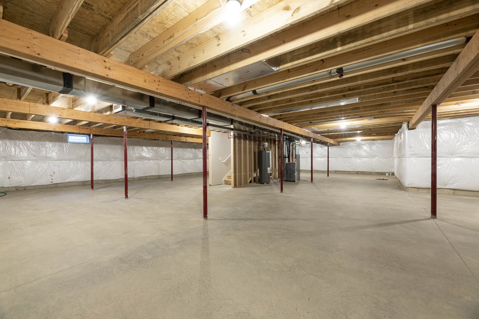 Basement room with exposed wood ceiling beams, visible metal pipes, concrete floor, red support poles, and unfinished walls.