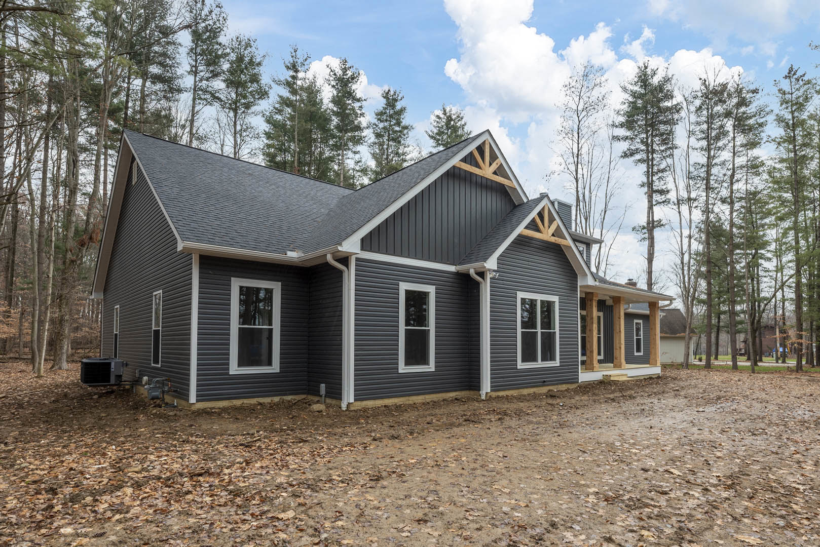 Partially built house with grey siding, white-framed window, black construction equipment on dirt ground, surrounded by trees under cloudy sky