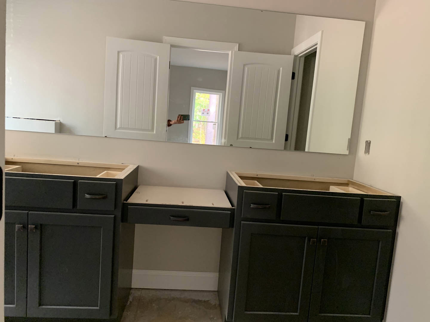 Modern bathroom featuring black cabinetry with drawers, white countertop, large wall mirror, window with signage, and a hand holding a cell phone near the sink.