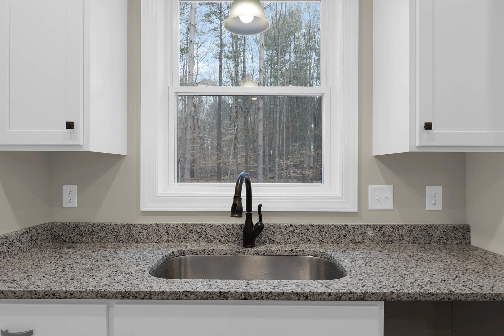 Granite countertop with stainless steel sink and modern faucet, white cabinetry, wall outlet, pendant light above, large window overlooking trees