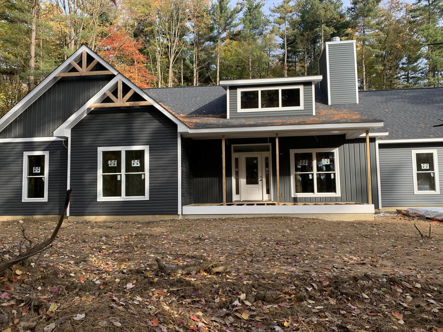 Two-story house with white-framed windows and glass-paneled white door, surrounded by tall trees and leafy ground, light-colored siding and cottage-style roof visible