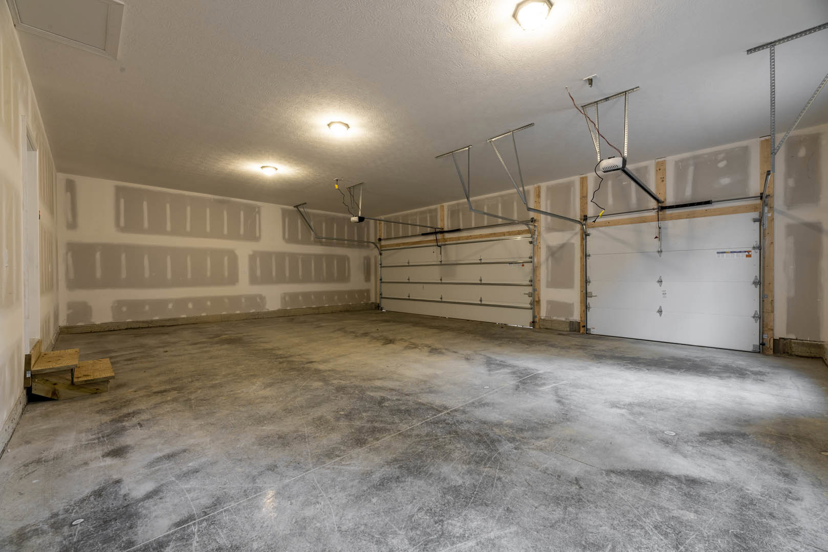 Garage interior with smooth concrete floor, white ceiling, overhead lighting, white garage door featuring metal hinges, and a wooden step stool near the wall.