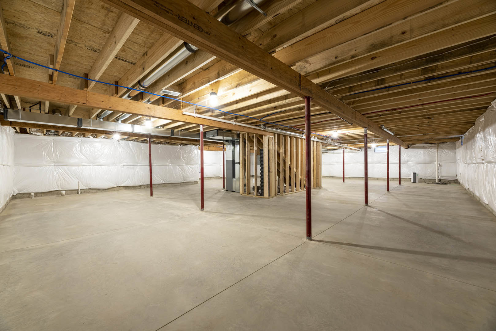 Exposed wooden ceiling beams, concrete floor with red support poles, white wall partially covered by large plastic sheet, visible pipes and construction materials in an unfinished