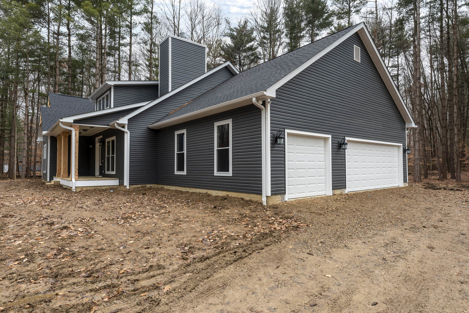 White siding house with black roof and chimney, white garage door, large window, dirt path with tire tracks, surrounded by trees.