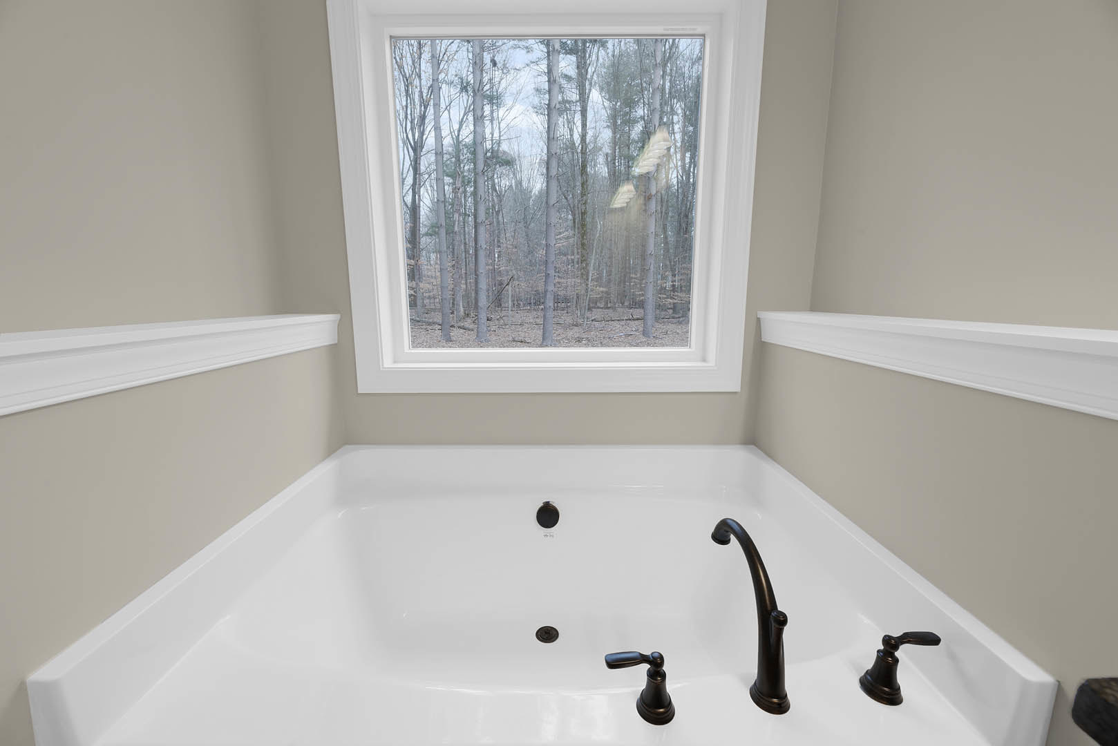Freestanding white bathtub beneath large window with view of green trees, black faucet, white wall molding, and black towel rack