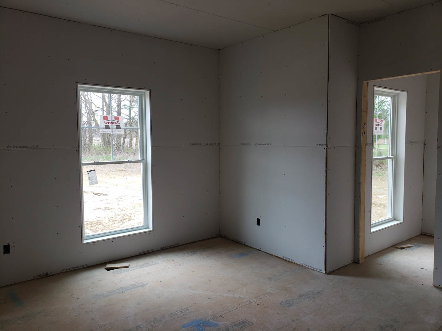 Bright room with white plaster walls, large windows overlooking a grassy yard, wood flooring, and a single door; window features a sign with red and black text.