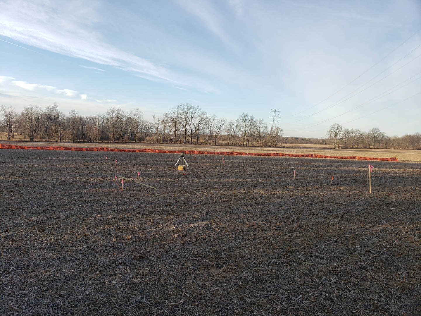 Grassy field with scattered orange poles and wires, tripod-mounted survey equipment, distant metal tower, cloudy sky overhead