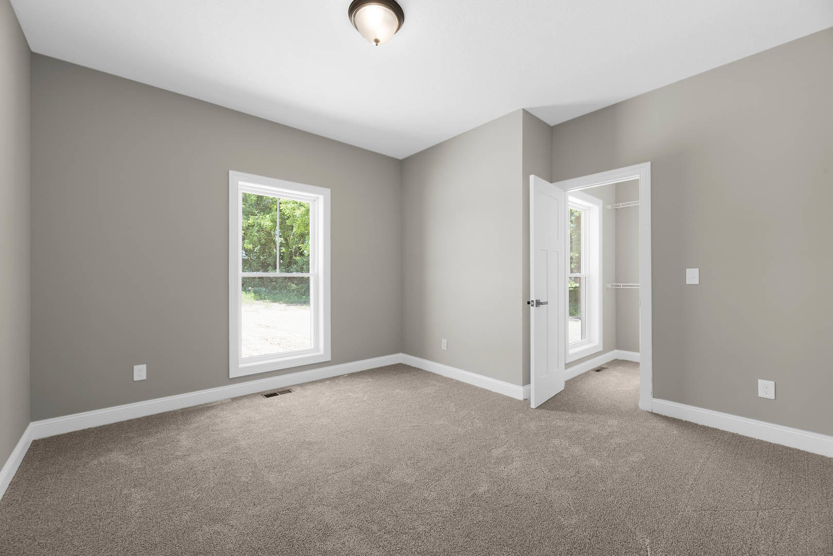 Bright room with white paneled door, large windows overlooking trees, neutral carpet flooring, modern ceiling light fixture, and crisp white walls with crown molding
