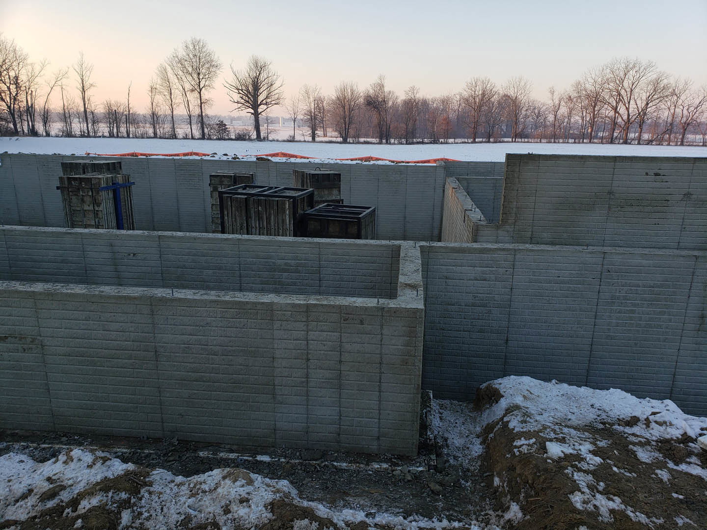 Snow-covered construction site bordered by leafless trees, stacks of wooden crates and concrete blocks visible in foreground