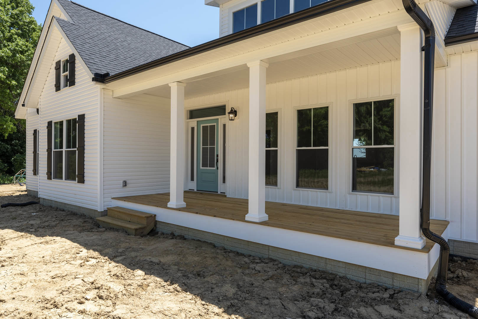 White siding exterior with green and blue front doors, wood porch and steps, large windows reflecting trees, shingled roof under clear sky