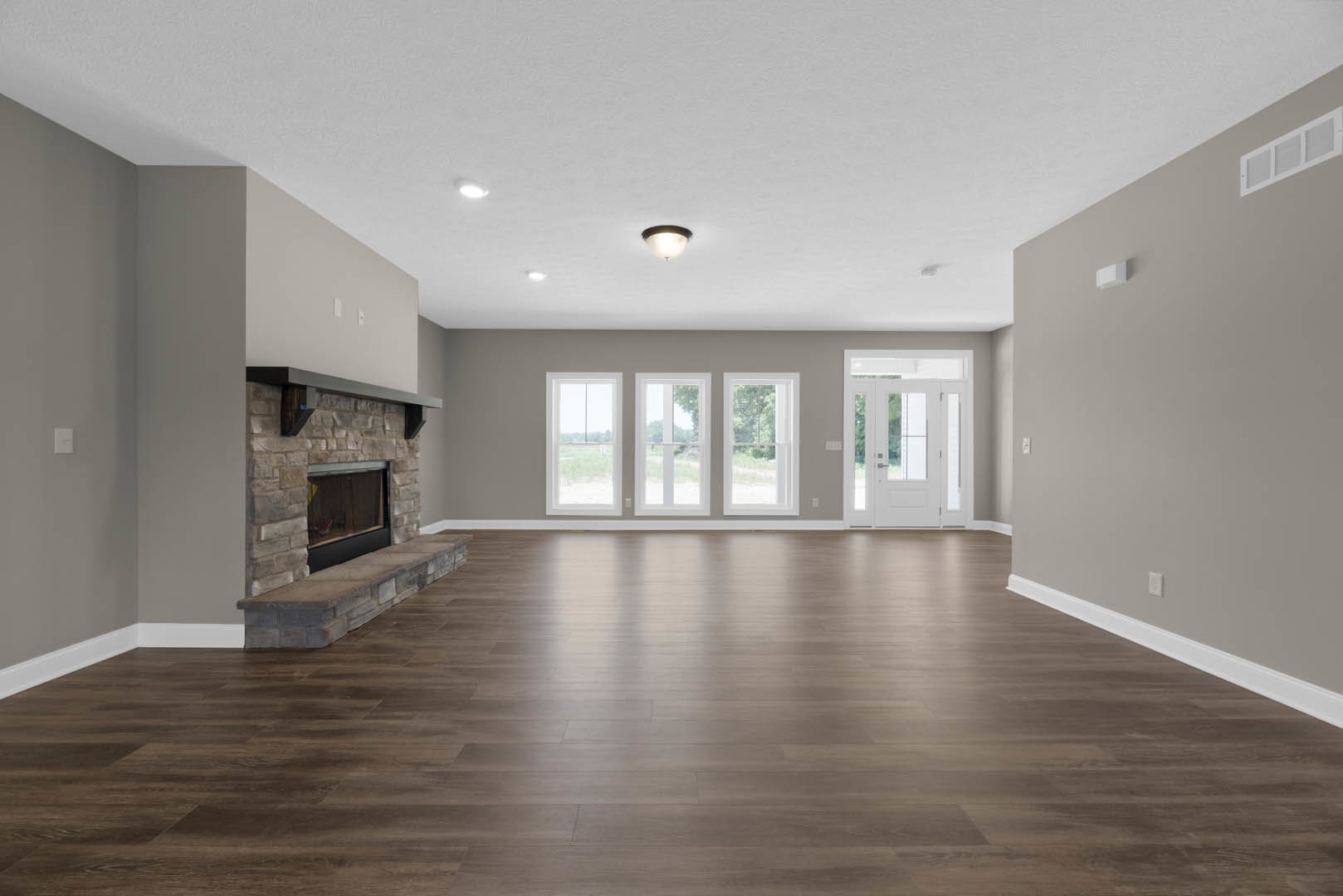 Living room with hardwood flooring, stone fireplace, ceiling light fixture, white door with glass panels, windows overlooking trees, plaster walls.