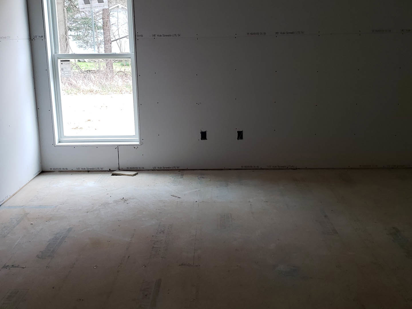 White-framed window set in plaster wall with visible holes, concrete floor featuring a piece of wood, close-up of white roll and screen in unfinished room